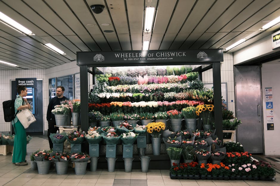A vibrant floral display at Wheeler of Chiswick within a train station, featuring a variety of fresh flowers arranged in tiered rows. The arrangement includes large bouquets of roses, tulips, daisies,