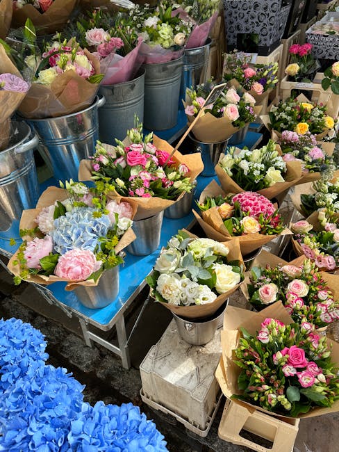 An outdoor display of various fresh flower bouquets arranged in metal buckets and wrapped in brown kraft paper, featuring pastel pink roses, white spray roses, pale green hydrangeas, and small yellow