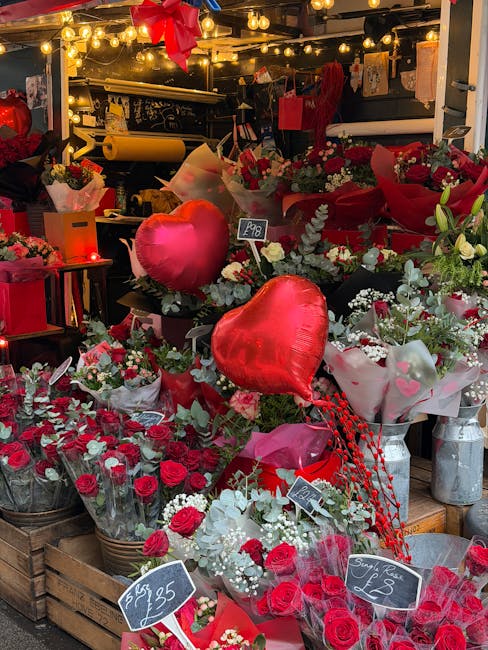 A vibrant display of floral arrangements and gifts at a flower stand, featuring lush bouquets of red roses, white lilies, and pink carnations, all wrapped in decorative paper and cellophane. The arran