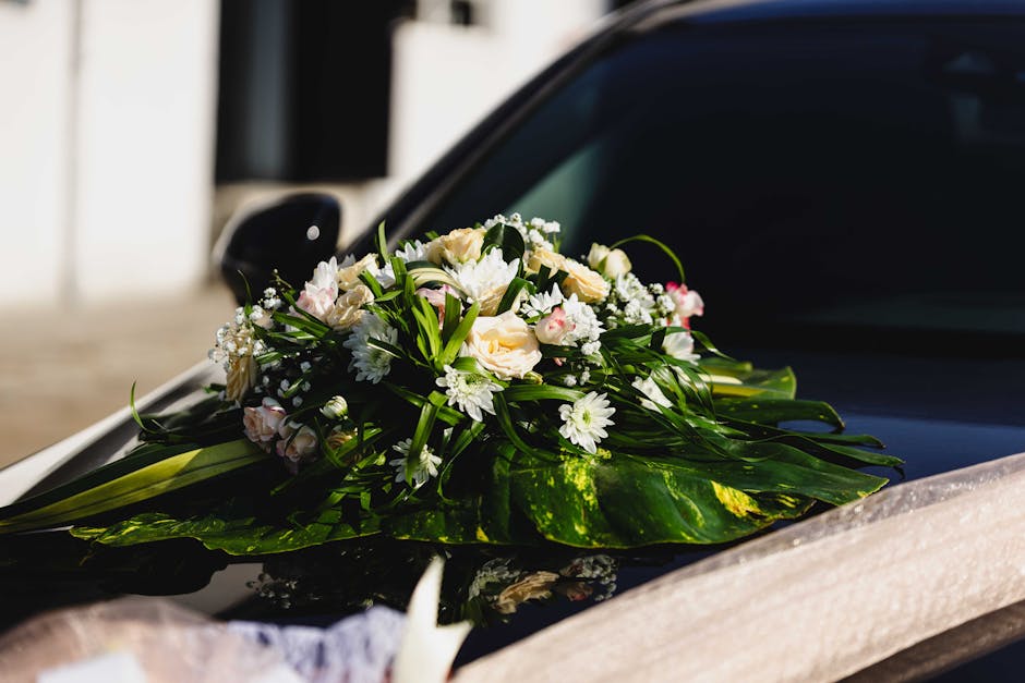 A delicate floral arrangement placed on the hood of a black car, featuring a bouquet of fresh white roses, small white daisies, and baby's breath, accented with lush green leaves and long stems, all w