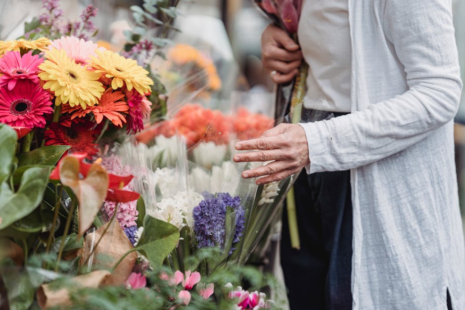 A person wearing a light grey cardigan and dark trousers is selecting fresh floral bouquets at a flower market, with their left hand gently touching a bouquet of vibrant pink, yellow, and red gerbera 