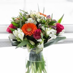 This artful Balham bouquet is an exploration of harmony and contrast, where structure and colour dance in perfect balance. Creamy, petal-rich gerberas create a gentle halo around bold orange centres that glow against the backdrop, their joyful disposition matching the lively rhythm of Balham's community. Flanking them, fuchsia-magenta roses spiral gracefully, adding a note of luxury and heartfelt emotion. The interior spaces are brightened by clusters of white alstroemeria and petite, ivory spray roses-multi-petaled and delicate as fresh morning dew-imbuing the whole with a feeling of lightness and vitality. Nestled at the base, broad, dark green aspidistra leaves wrap protectively, their glossy texture contrasting sharply yet beautifully with the softness above-much like the diverse character found along Bedford Hill. Sprigs of additional green foliage weave through the bouquet, building both fullness and dynamic texture. Enclosed in a slender glass vase, the arrangement is softly illuminated from behind, ensuring the vivid colours and layered beauty remain the visual focus. The effect is one of optimism and generous spirit-a floral union perfect for gathering friends, marking new chapters, or simply bringing a flourish of Balham's charm into your home.